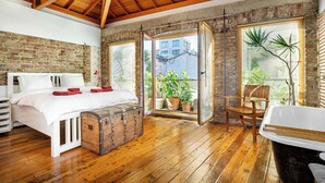 Ein Schlafzimmer mit Holzbalkendecke, einer Ziegelwand, einem Bett mit weißer Bettwäsche, einer Holzablage und Blick ins Freie durch ein Fenster.