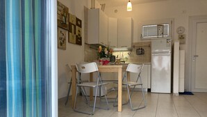 A compact kitchen with white cabinets, a small dining table with chairs, a refrigerator, and a window with blinds. - Modern Flat Just 150 mt From the sea (Rosolina)