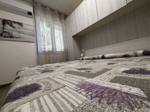 A bedroom with a bed, a window with sheer curtains, a wall-mounted air conditioner, and a painting of a beach scene. - Accommodation in the Vibrant Heart of Rosolina (Rosolina)