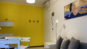 Una stanza con una parete gialla, un divano grigio con cuscini, una scrivania bianca con sedie e un quadro appeso al muro.