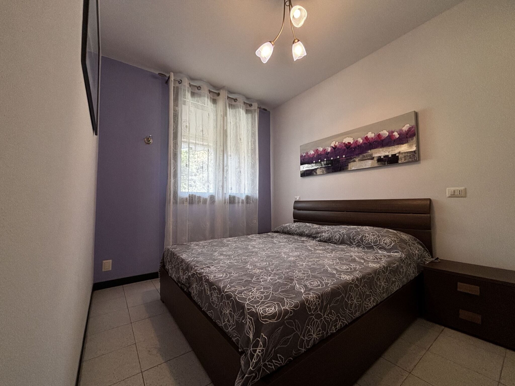Un dormitorio con cama, cortinas, un cuadro y una mesita de noche.