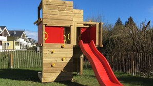Une structure de jeu en bois, dotée d’un toboggan rouge et d’échelons d’escalade, installée dans un espace verdoyant, avec des maisons en arrière-plan.