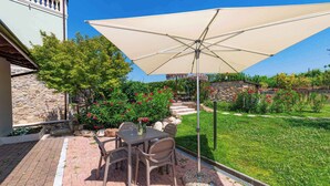 Un patio al aire libre con una mesa y sillas bajo un gran paraguas, rodeado de un jardín cuidado y un muro de piedra.