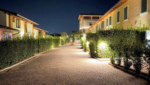 Un camino pavimentado flanqueado por edificios con iluminación exterior y bordeado de setos.