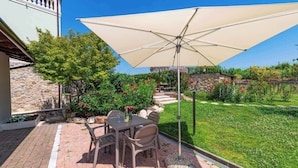 Un patio al aire libre con una mesa y sillas bajo un gran paraguas, rodeado de un jardín cuidado y un muro de piedra.