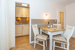 A dining area with a wooden table and white chairs, a kitchen with white cabinets and a countertop, and a wall-mounted light fixture. - Dünenburg Apartment 15 - Holiday apartment with balcony near the beach in the heart of (Sylt)