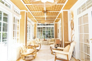 Une piĂšce lumineuse, meublĂ©e de mobilier en osier, dotĂ©e de grandes fenĂȘtres et dâun ventilateur de plafond.