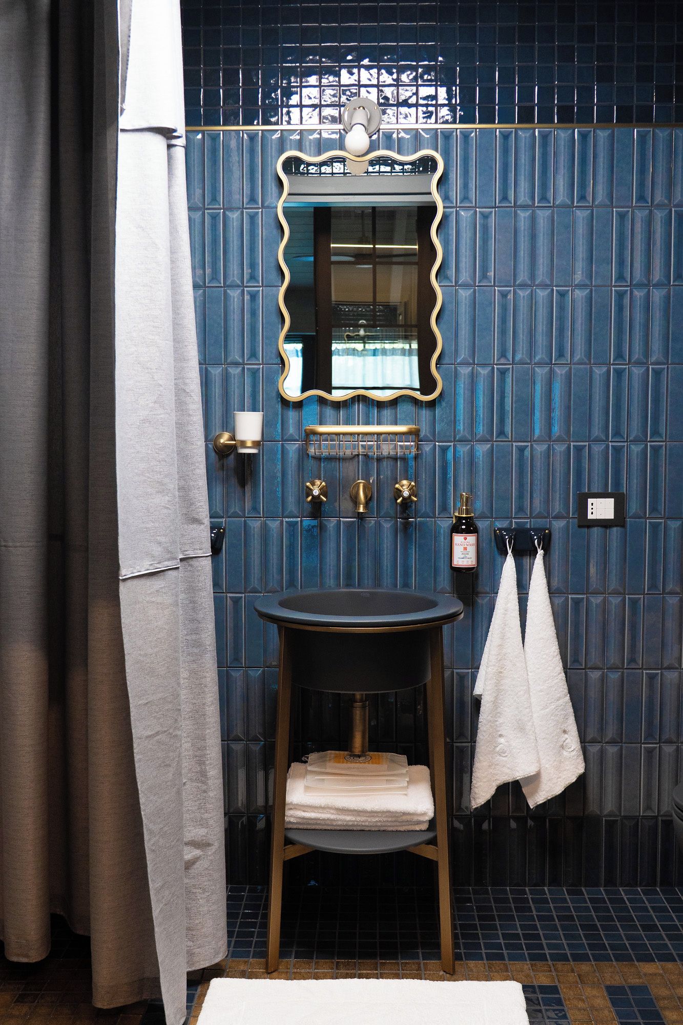 Ein Badezimmer mit blauen Fliesen, einem runden Waschbecken, einem Spiegel mit goldener Rahmung und zwei Handtüchern an Haken.