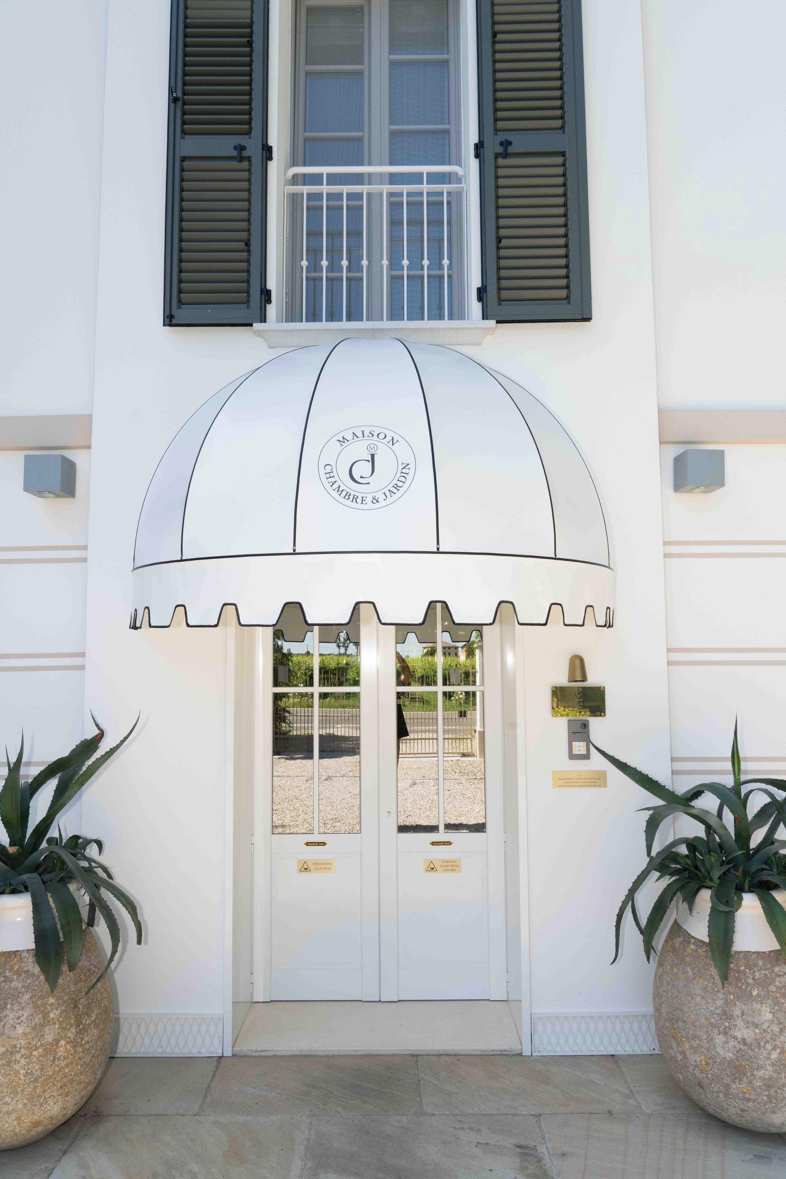 A hotel entrance with a white facade, a large arched awning, and two potted plants on either side.