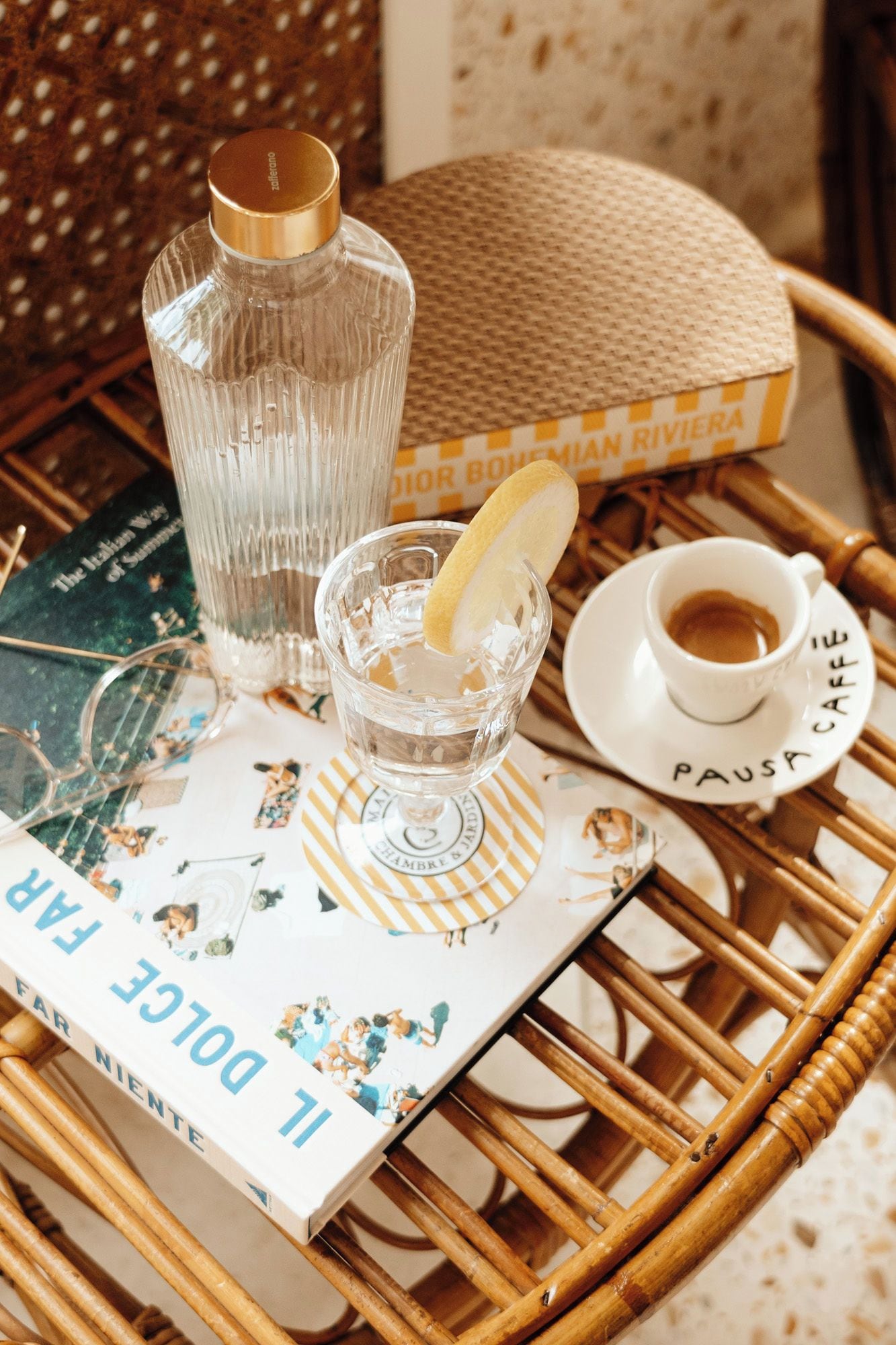A wicker table with a book, a glass of water with a lemon slice, a glass of coffee, and a bottle of water.
