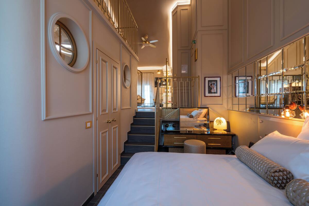 Une chambre d’hôtel avec un lit, un bureau doté d’une lampe, un miroir et un escalier menant à un étage supérieur.