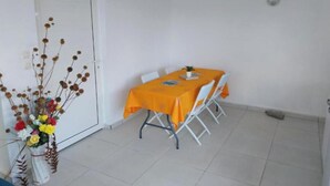 Ein Essbereich mit einem Tisch, der mit einer orangefarbenen Tischdecke bedeckt ist, weißen Stühlen und einer Blumenvase auf der linken Seite.