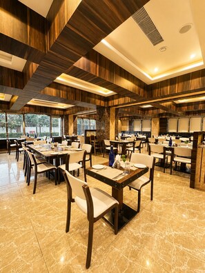 A spacious restaurant with multiple dining tables and chairs, a high ceiling with wooden paneling, and large windows allowing natural light. - Golden Tulip Kukas Jaipur (Amer)