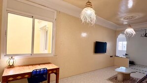 Una habitación amplia con un escritorio de madera, una silla azul, un televisor y una lámpara decorativa en el techo.