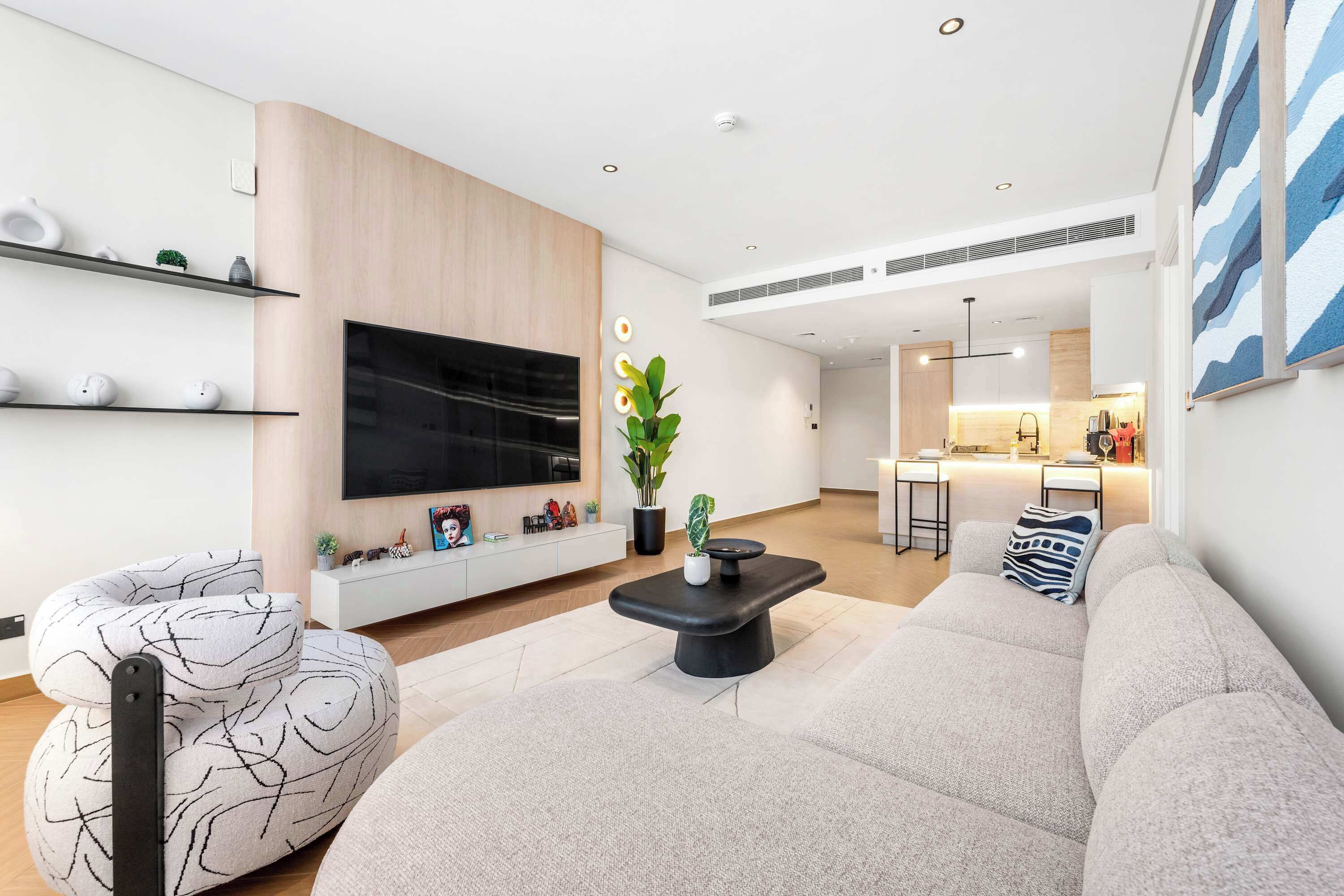 Uma sala de estar moderna com um sofá, uma mesa de centro e uma televisão. Uma cozinha visível ao fundo.