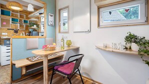 Eine gemütliche Küche mit Holzschränken, einem Esstisch und einem Stuhl. Ein Fenster mit herzförmigen Verzierungen und ein Regal mit Küchenutensilien.