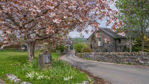 Une maison en pierre avec une porte verte, entourée d’un mur de pierre et d’un espace verdoyant où fleurissent de magnifiques fleurs.