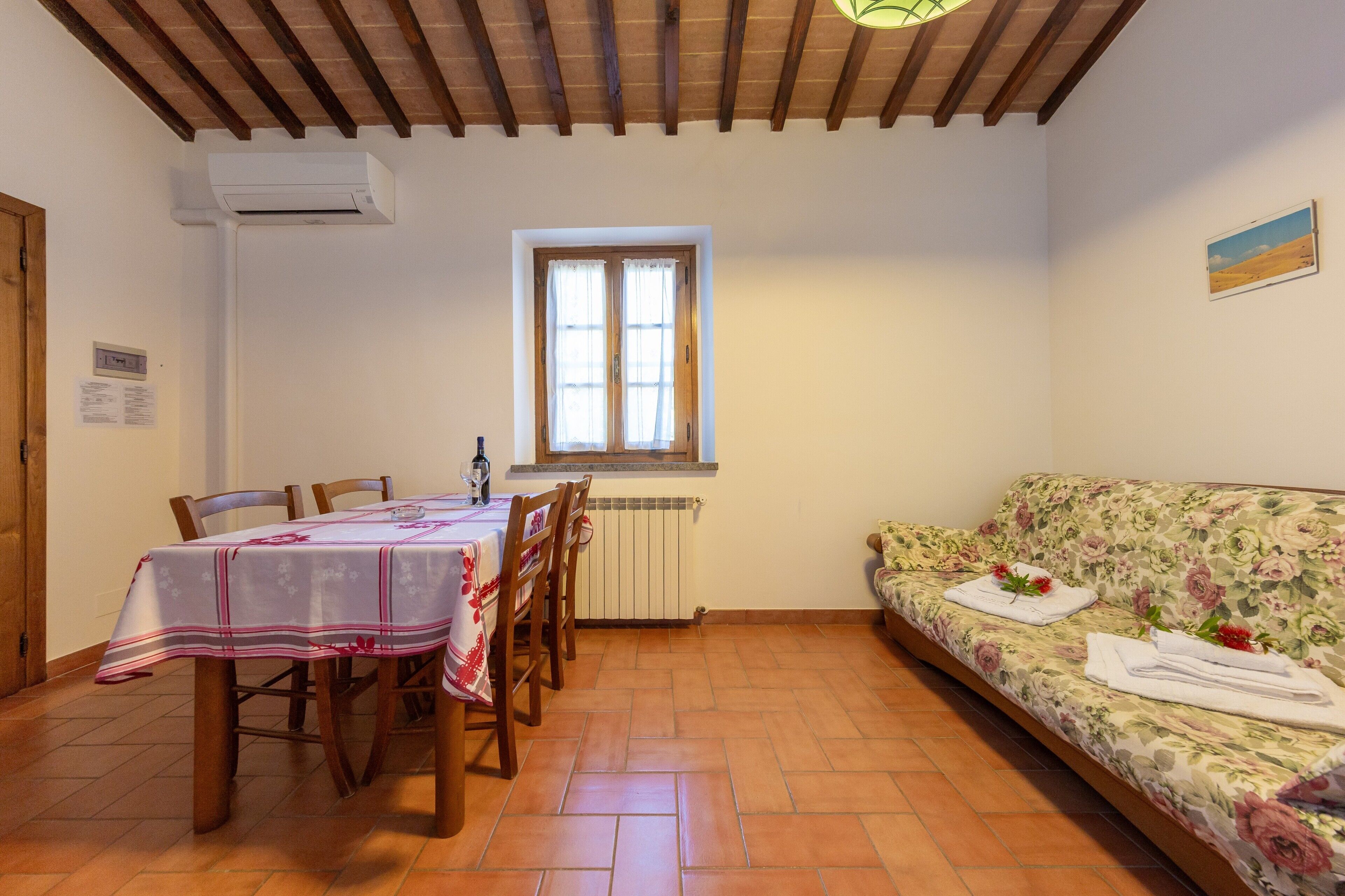 Un'area pranzo con tavolo apparecchiato per quattro, un divano con asciugamani e fiori, una finestra con tende e un soffitto in legno.