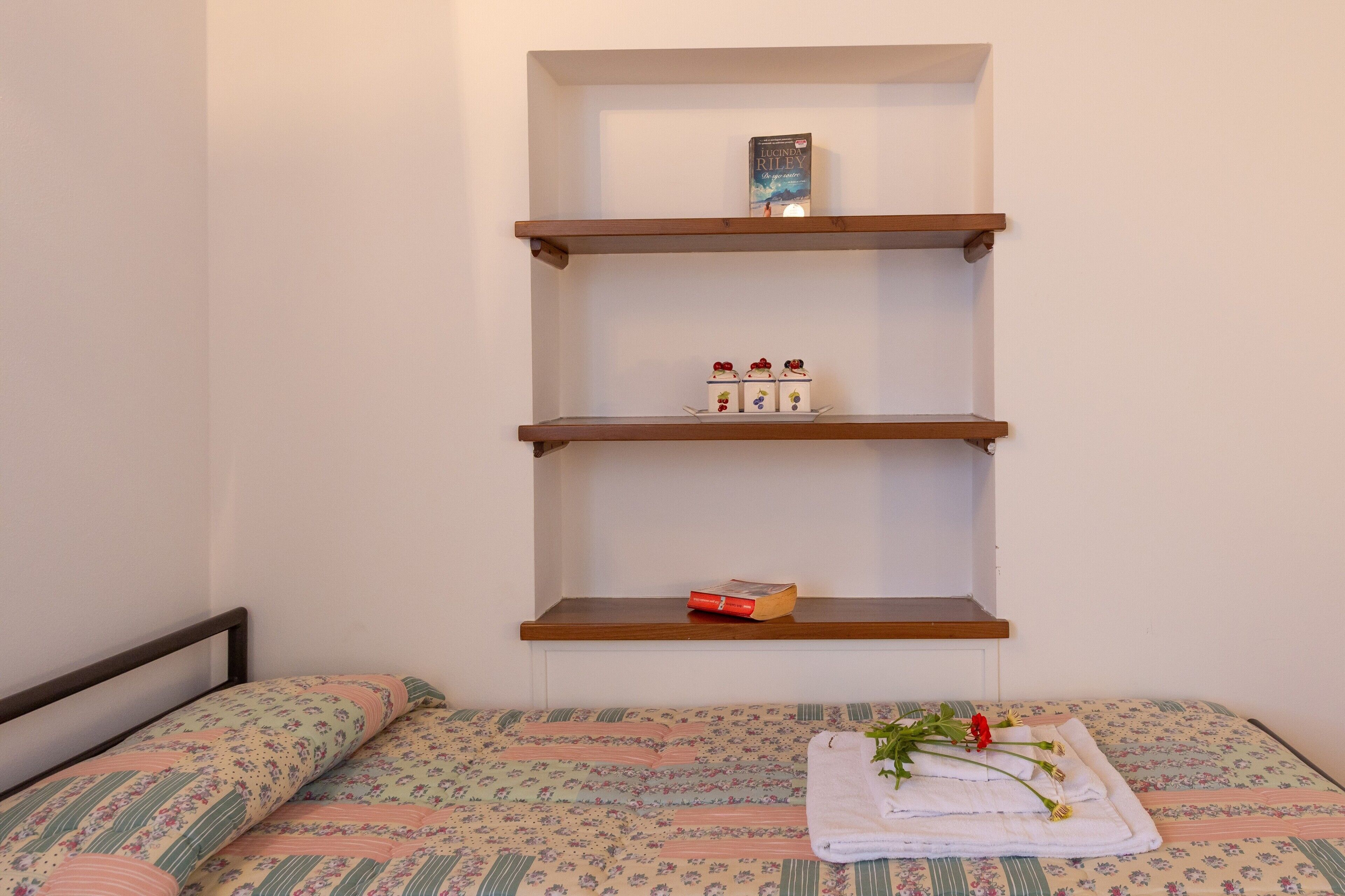 Une chambre avec un lit, une étagère fixée au mur où sont rangés des livres et une petite boîte, et une serviette soigneusement pliée, ornée de motifs floraux, posée sur le lit.