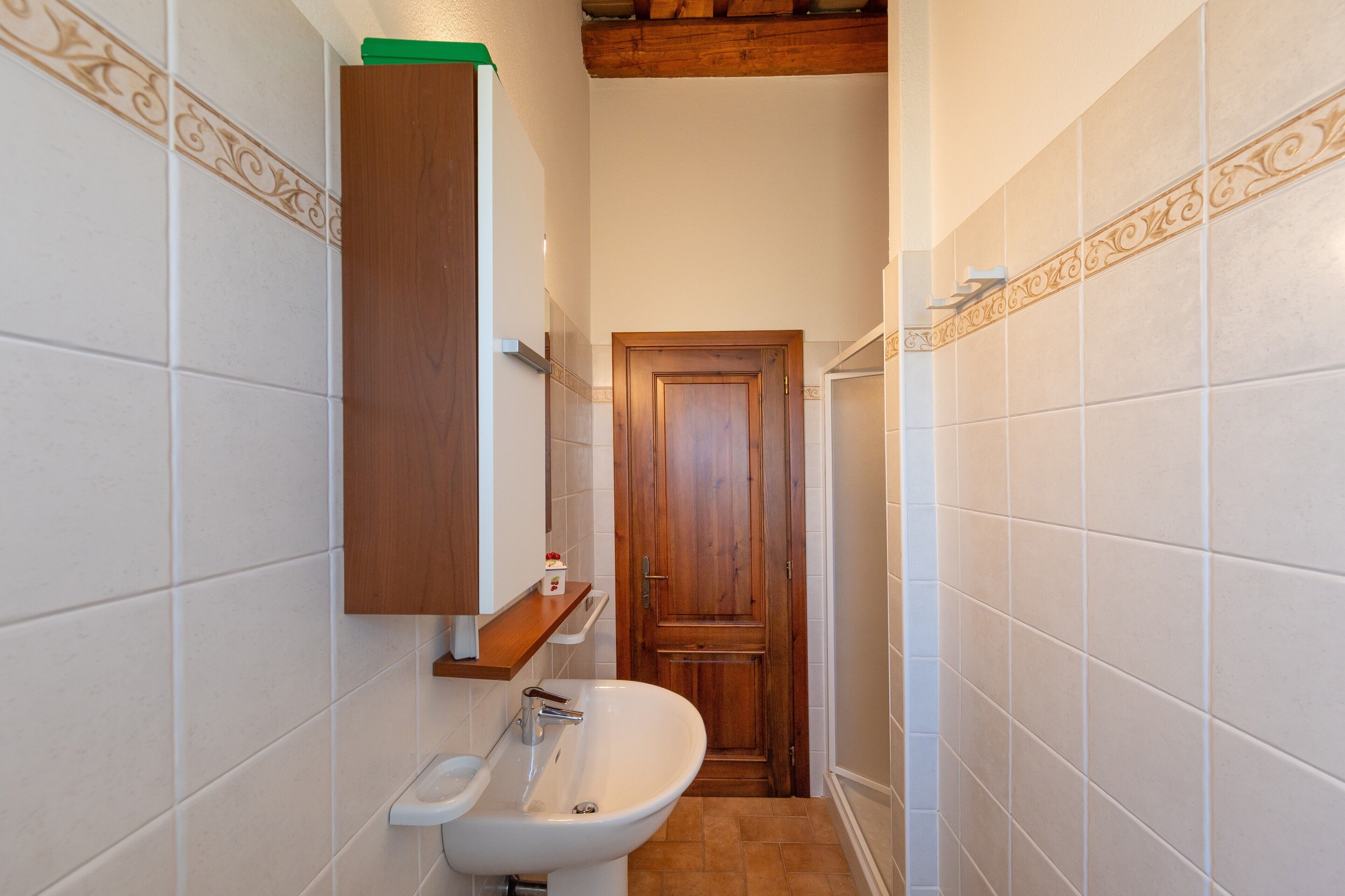 Un baño estrecho con lavamanos blanco, puerta de madera y estante sobre el lavamanos.