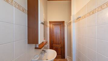 Un baño estrecho con lavamanos blanco, puerta de madera y estante sobre el lavamanos.