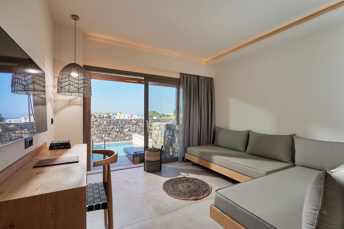 A modern living room with a sofa, a wooden desk, and a large window offering a view of a pool and cityscape.