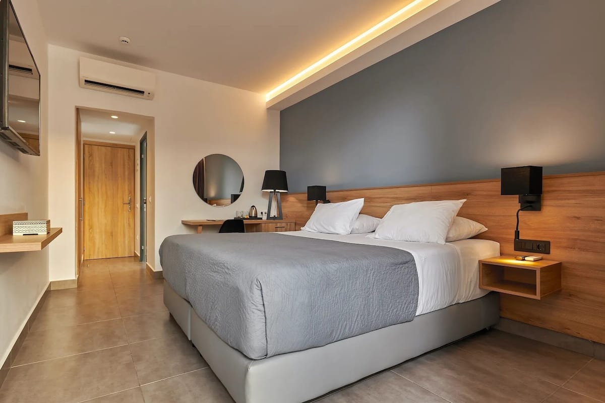 A modern hotel room with a large bed, bedside tables, a round mirror, and a wooden headboard.