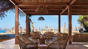 A covered outdoor seating area with wicker chairs and a central table, overlooking a beach with umbrellas and lounge chairs. - Sentido Amounda Bay (Malevizi)
