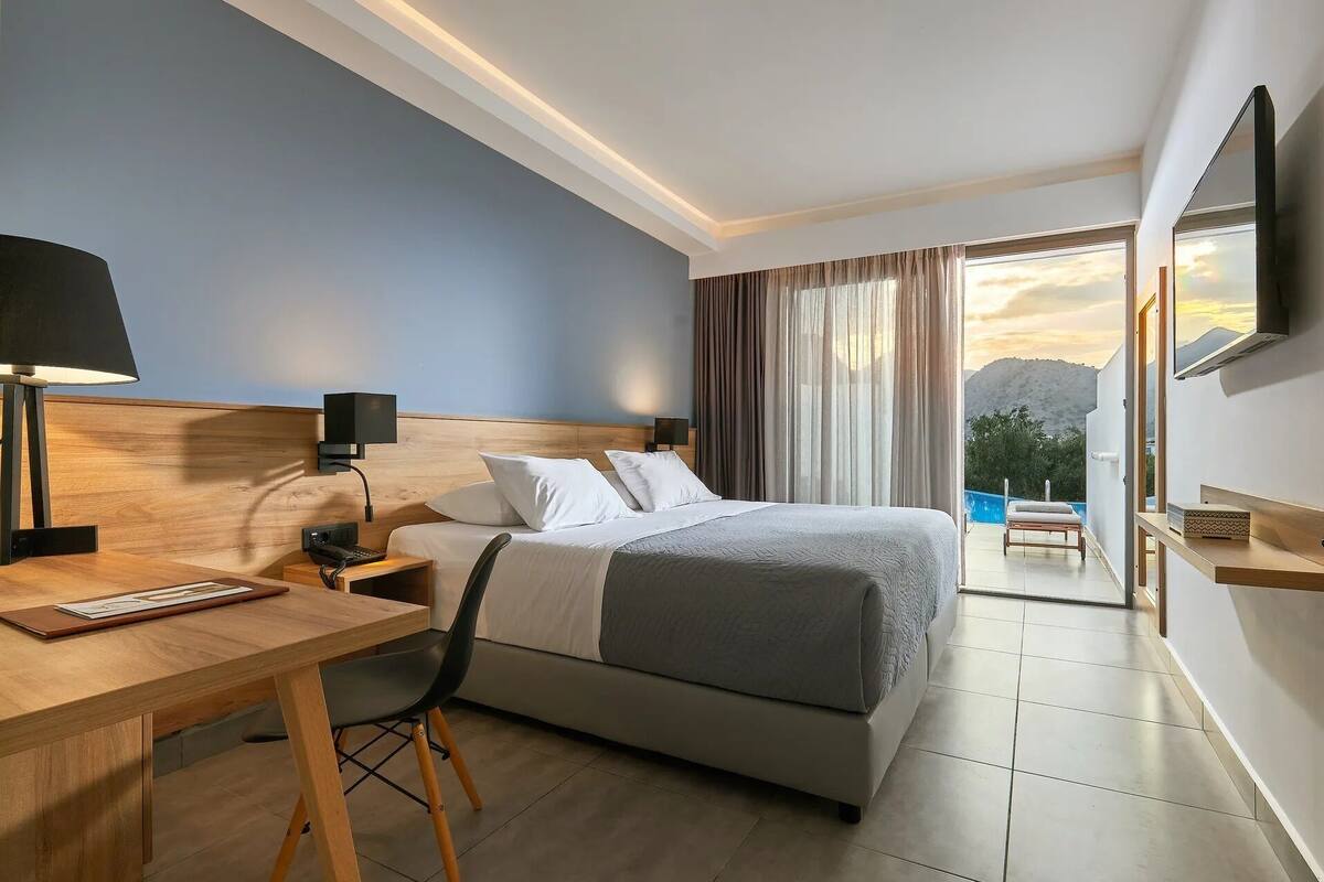A hotel room with a bed, a desk, a chair, a TV, and a balcony with a view of the mountains.