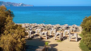 A beach resort with rows of sunbeds and umbrellas, a clear view of the sea, and a mountainous backdrop. - Sentido Amounda Bay (Malevizi)