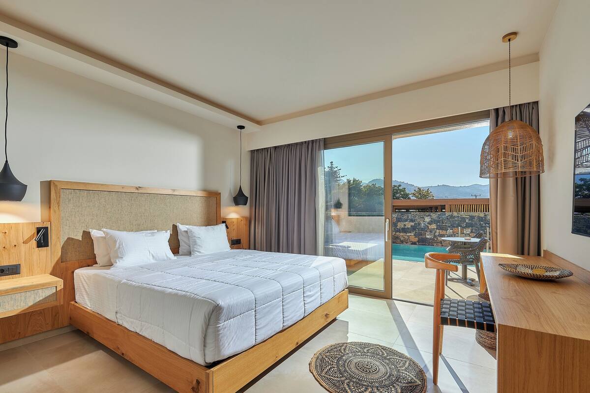 A modern bedroom with a large bed, wooden headboard, and a view of a pool and mountains.