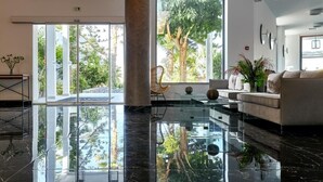 A modern hotel lobby with a glass floor, a sofa, a chair, and a large window offering a view of trees and a building. - Sentido Amounda Bay (Malevizi)