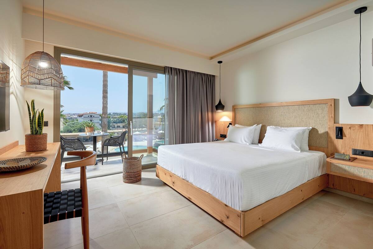 A modern hotel room with a large bed, a desk with a chair, a dining area with a table and chairs, and a view of the outdoors.