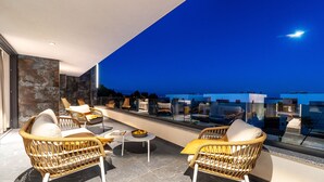A modern outdoor seating area with wicker chairs and a small table, overlooking a cityscape at dusk.