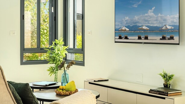 A room with a large window, a wall-mounted TV, a small table with a laptop, a vase with plants, and a bookshelf. - MAMAD - Tranquil Galilee Retreat by Sea N' Rent (Merom HaGalil)