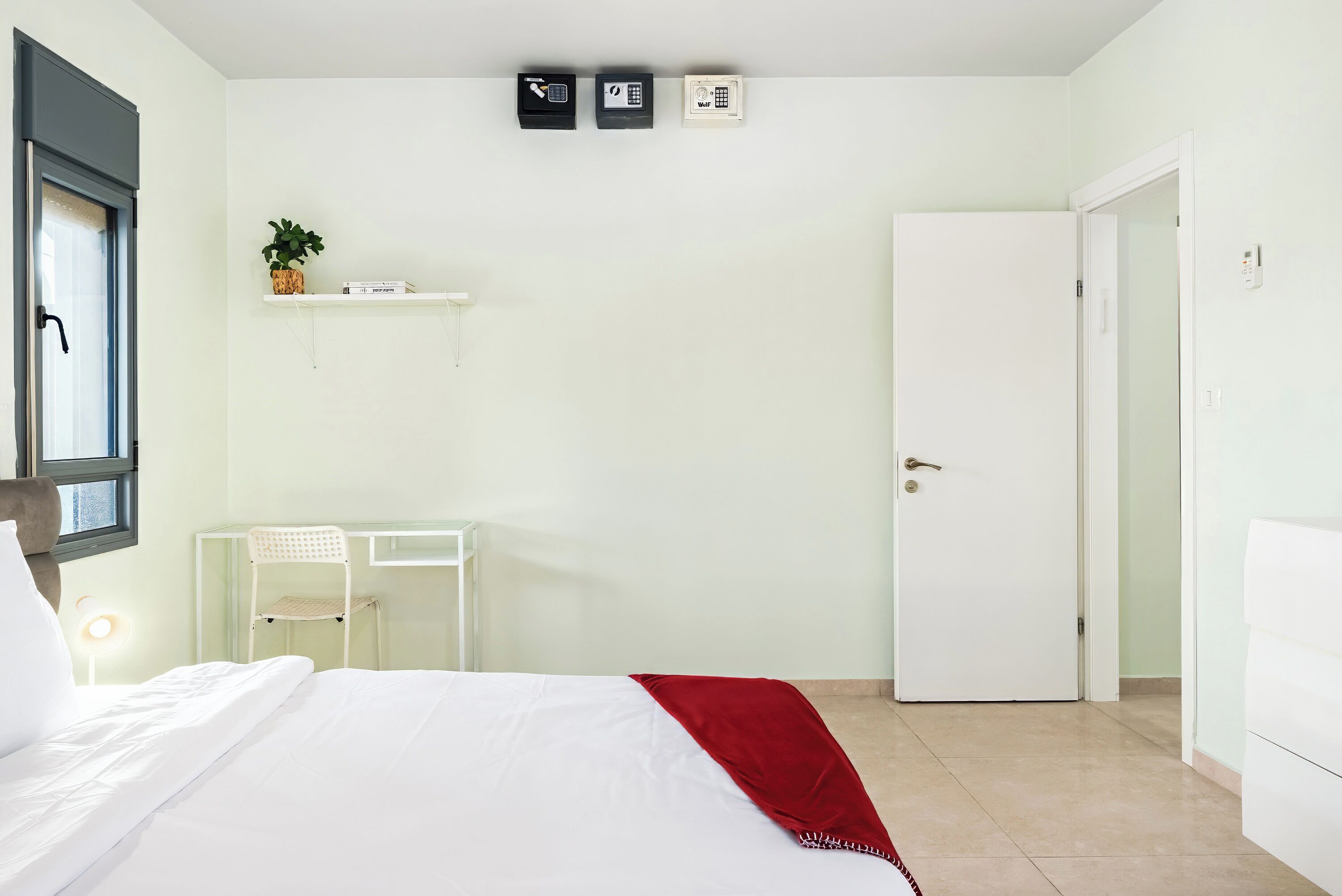 A bedroom with a bed, a desk, a chair, a door, and a wall-mounted phone.