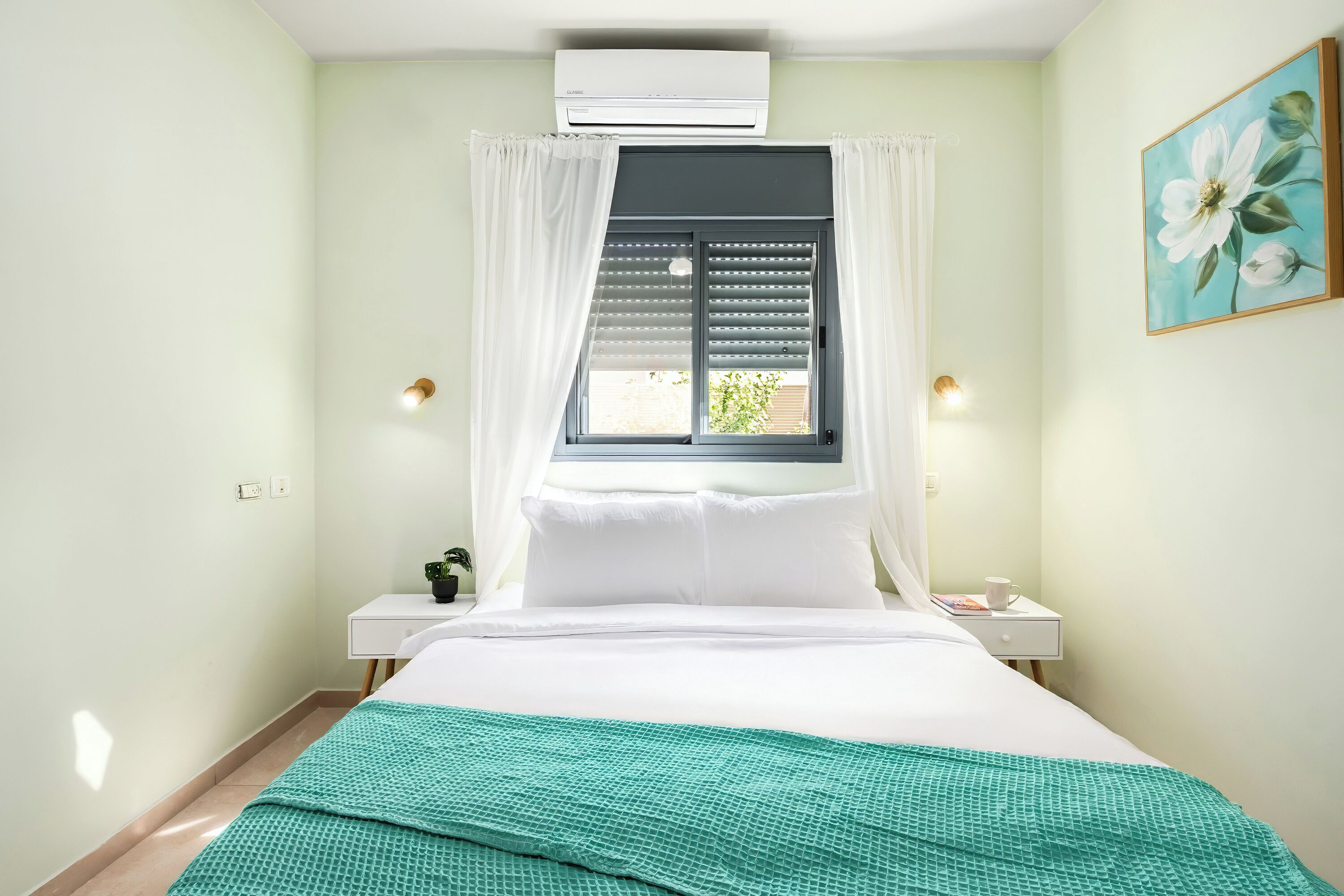 A bedroom with a bed, a window with curtains, a bedside table with a lamp, and a wall-mounted air conditioner.