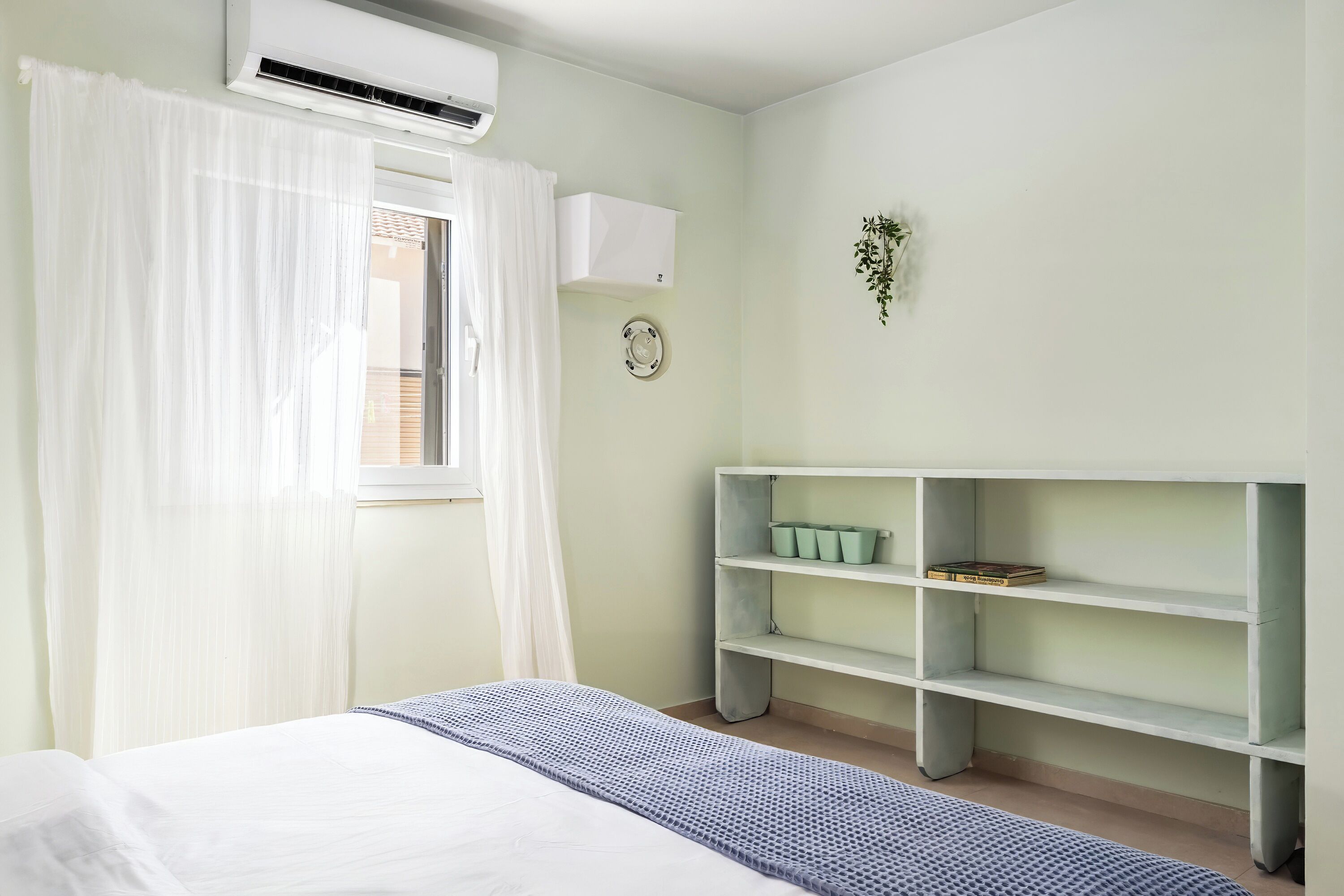 Un dormitorio con cama, cortinas blancas, unidad de aire acondicionado, estante en la pared con libros y un pequeño plato decorativo.
