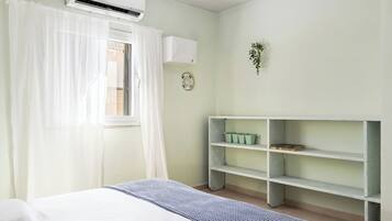 Un dormitorio con cama, cortinas blancas, unidad de aire acondicionado, estante en la pared con libros y un pequeño plato decorativo.