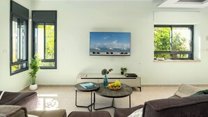 A modern living room with a large window, a painting, a small table with a vase and fruit, and a sofa. - MAMAD - Tranquil Galilee Retreat by Sea N' Rent (Merom HaGalil)