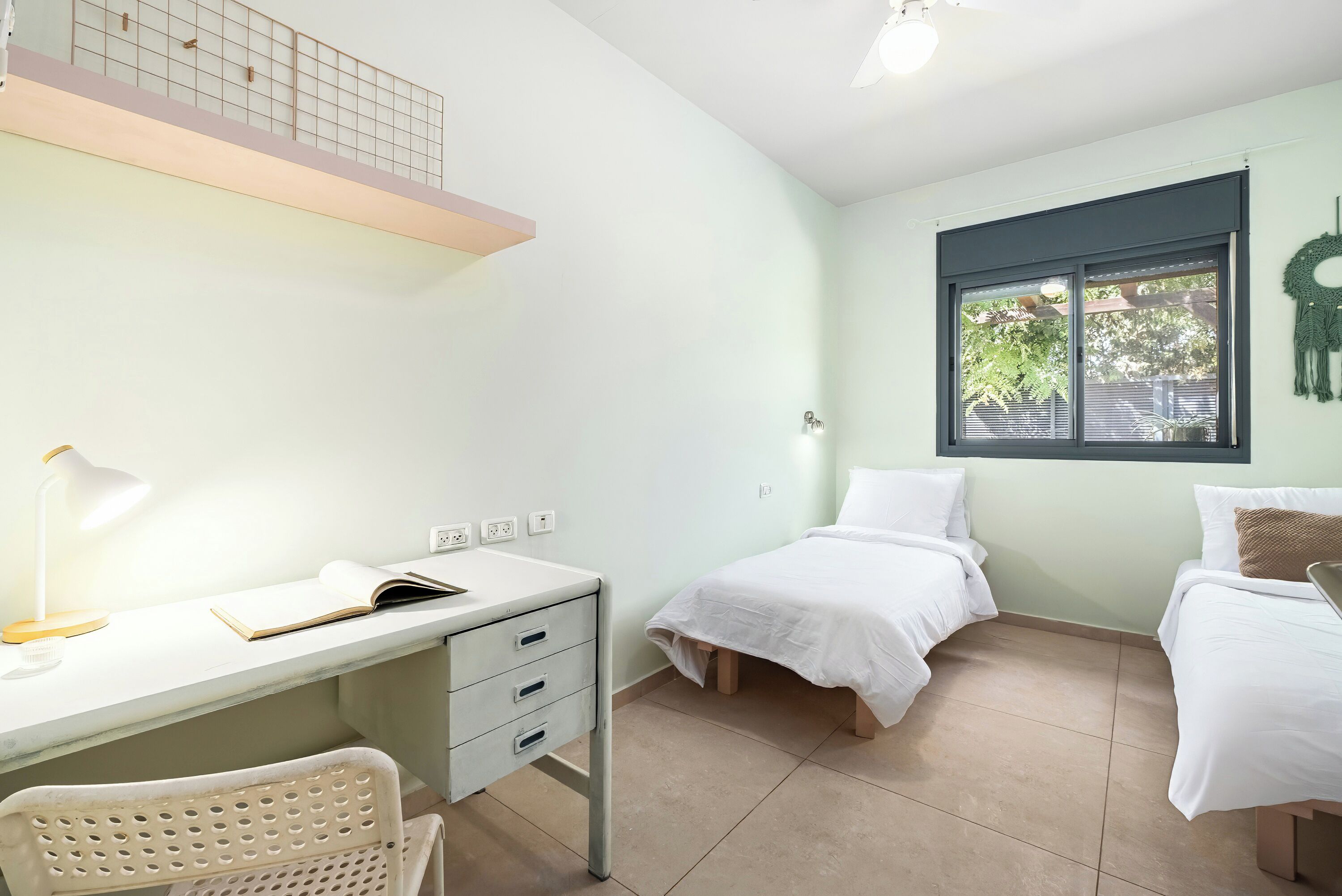 A bedroom with a desk, two beds, a chair, and a window with a view of trees.
