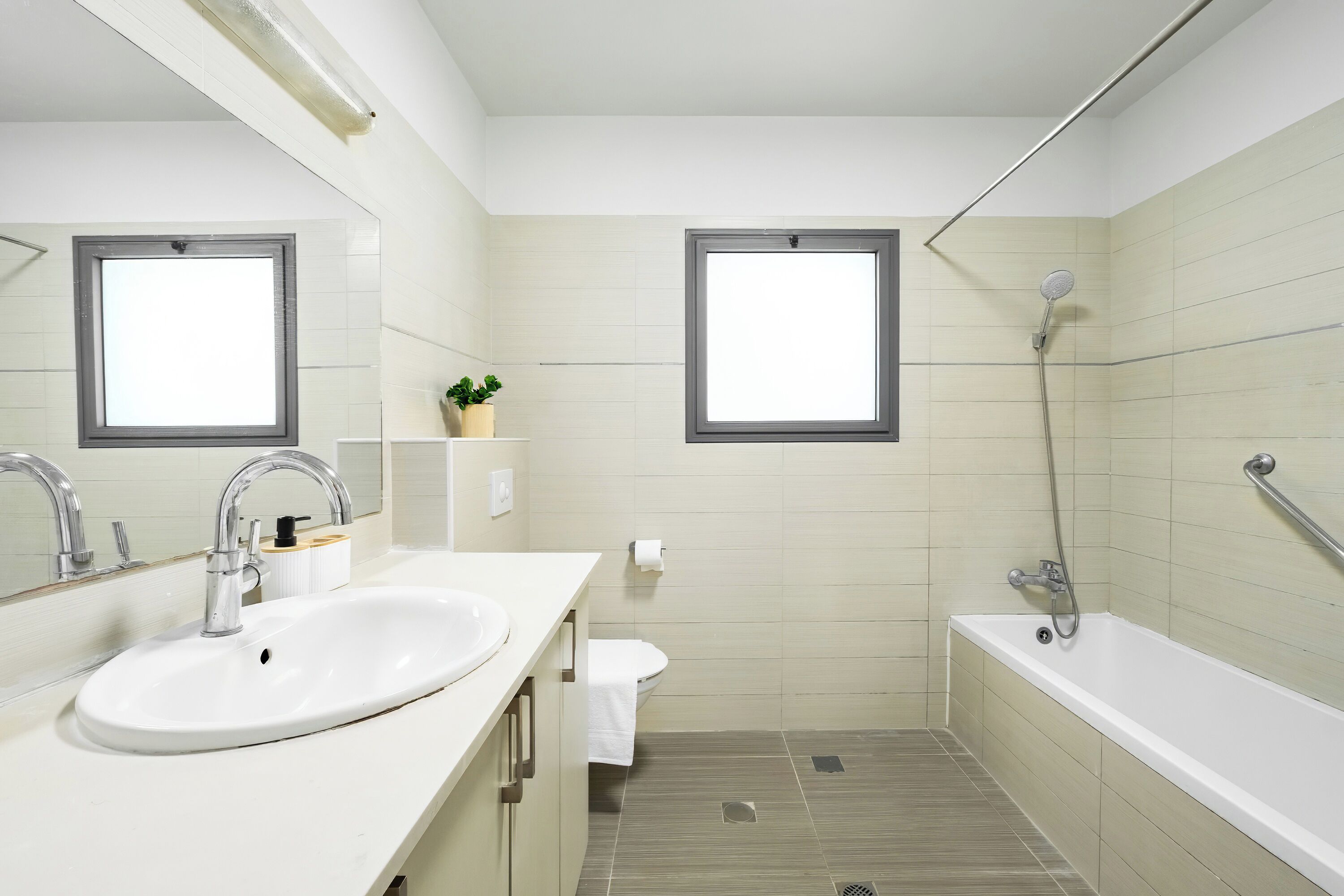 A modern bathroom with a white sink, a toilet, a shower, and two windows.