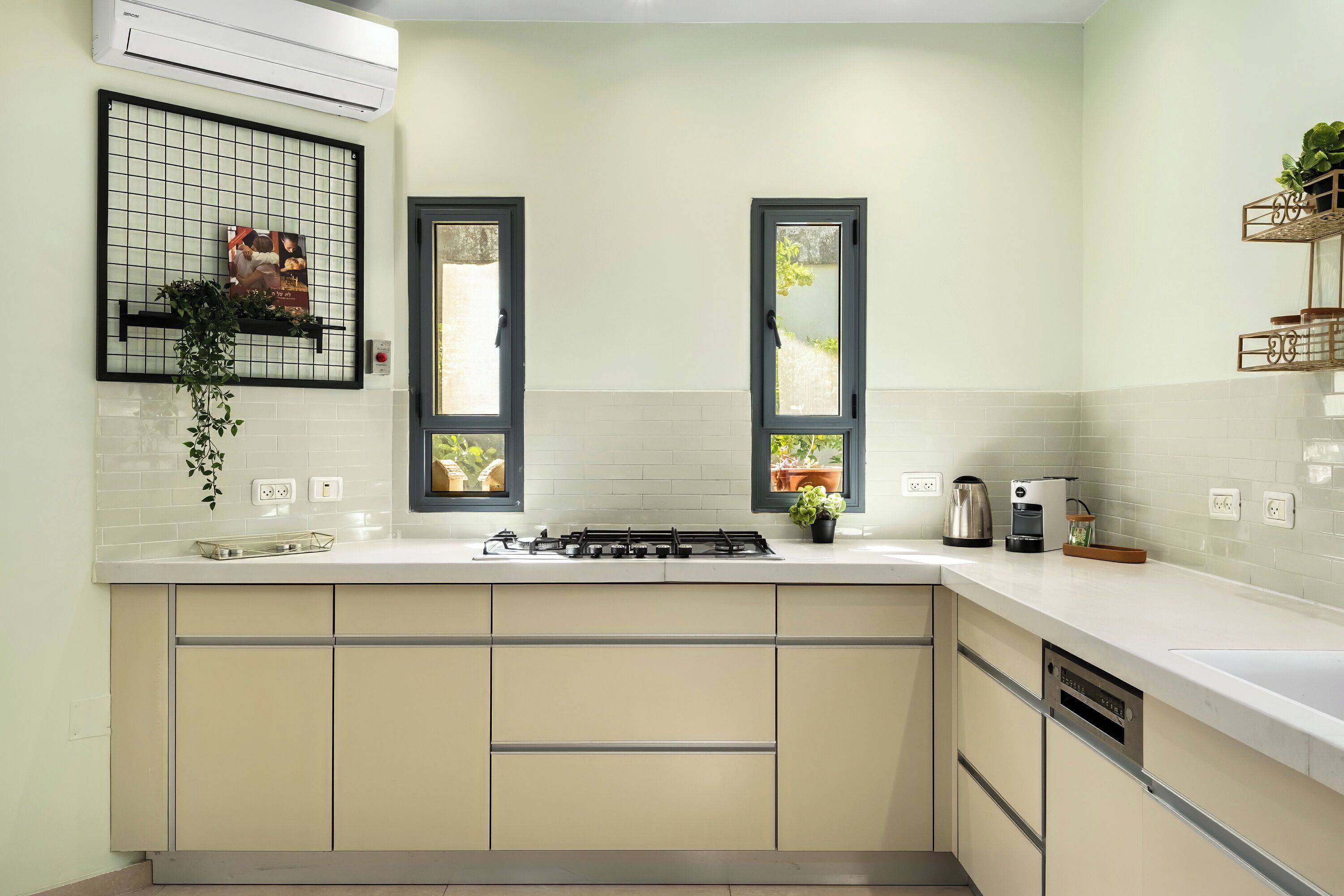 A modern kitchen with a gas stove, white countertops, beige cabinets, and two windows with black frames.