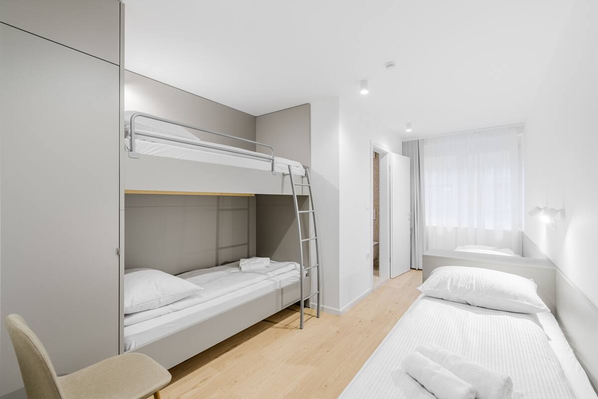 Une chambre d’hôtel moderne équipée d’un lit superposé, d’un lit simple, d’une chaise et d’une fenêtre avec des rideaux.