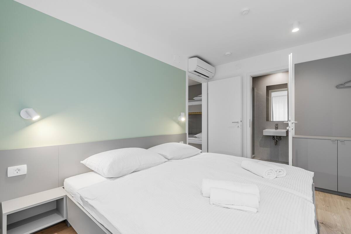 Une chambre d’hôtel moderne avec un lit bien fait, une table de chevet, une armoire intégrée et une salle de bain équipée d’un lavabo et d’un miroir.
