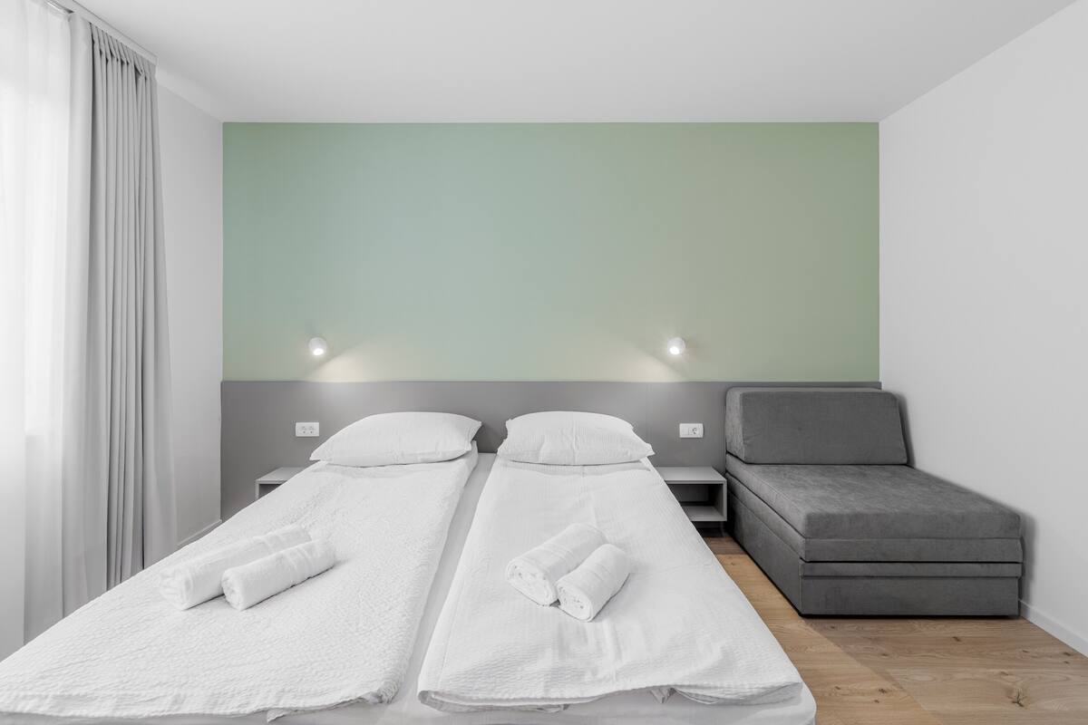 Une chambre double avec deux lits, un canapé gris et un mur de couleur vert clair.