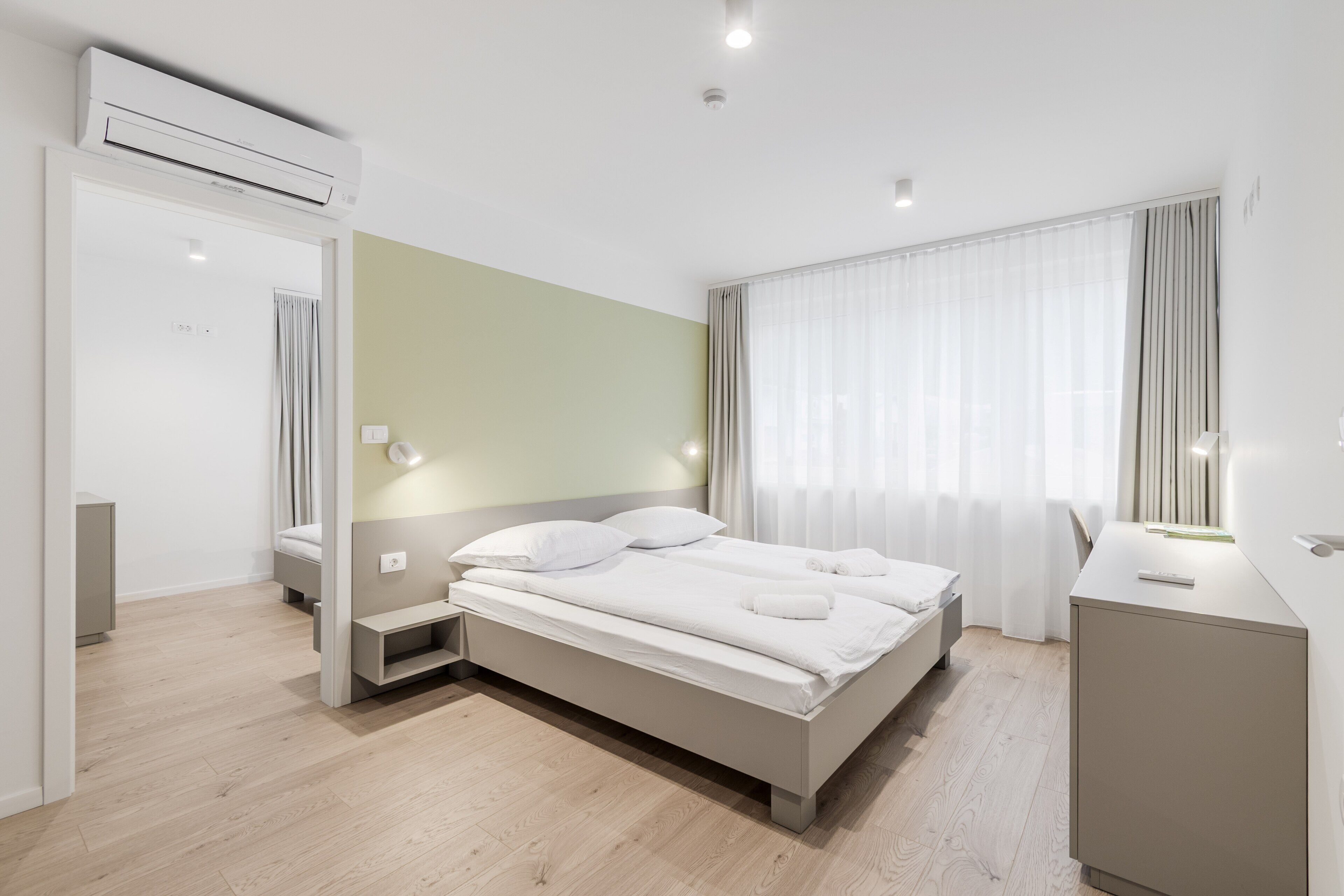 A modern hotel room with a large bed, bedside tables, an air conditioning unit, and a window with curtains.