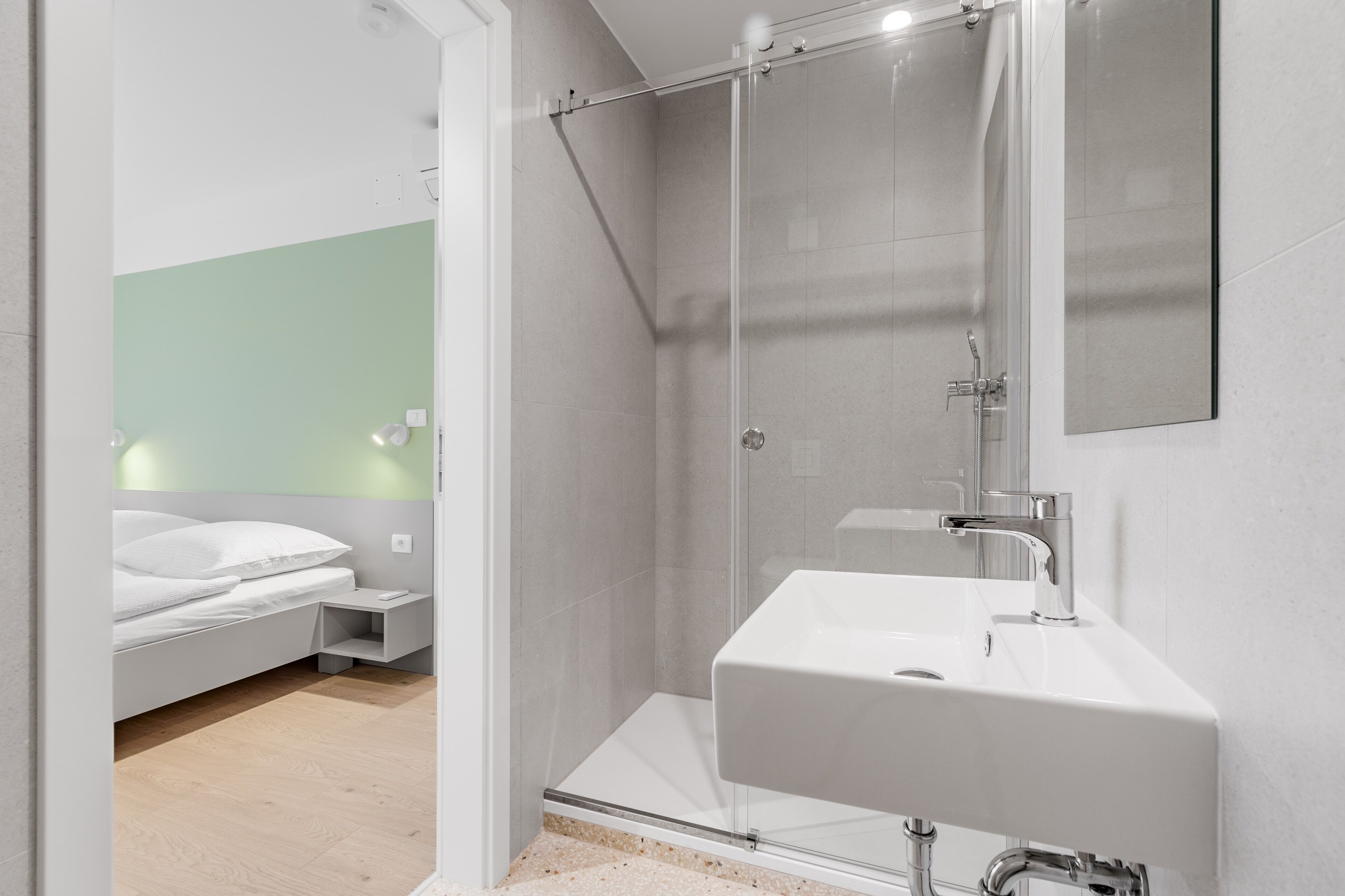A modern bathroom with a glass shower enclosure, a white sink, and a bedside table with a lamp.