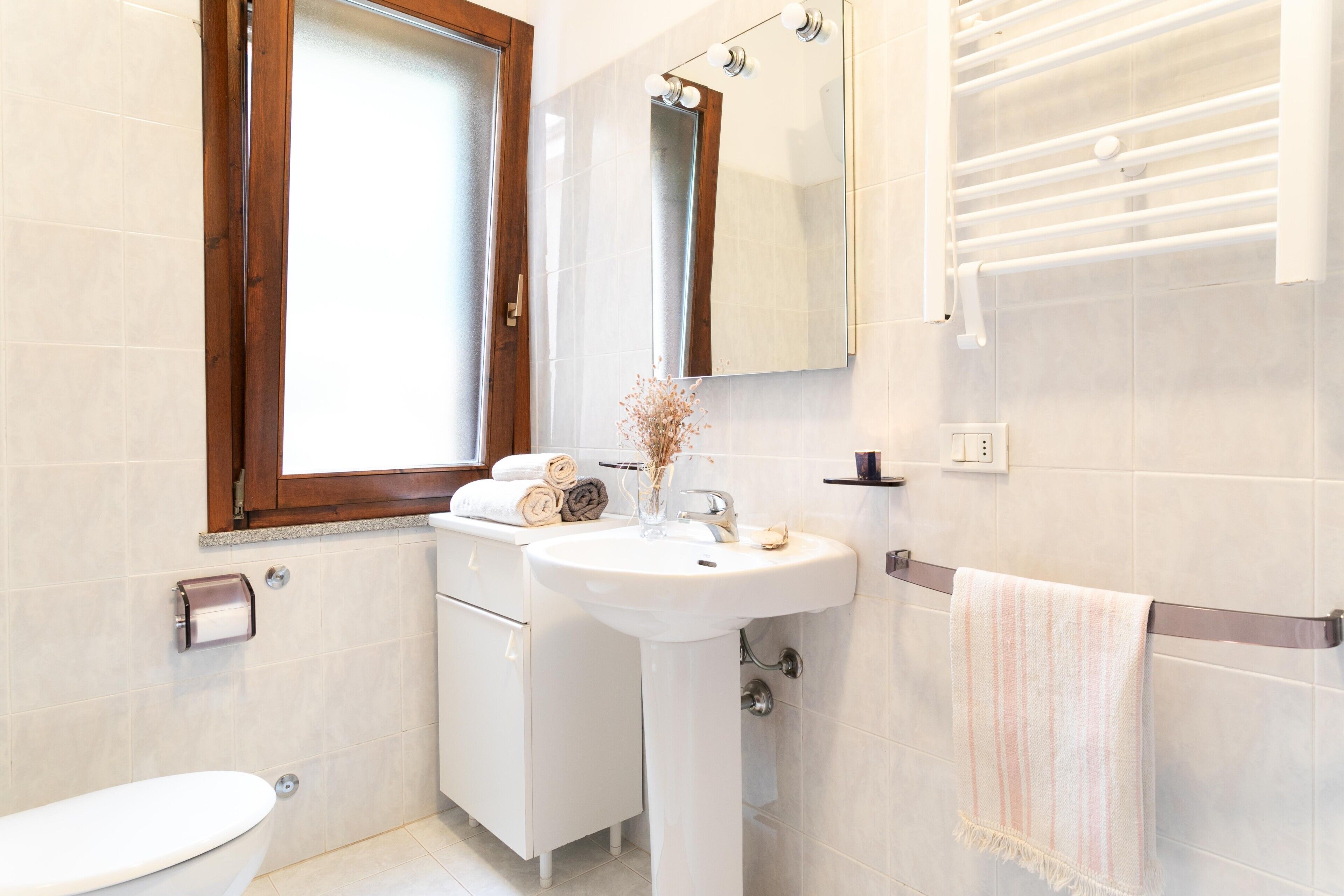 A bathroom with a toilet, sink, mirror, towel rack, and a window with a wooden frame.