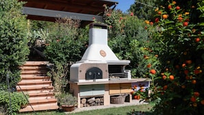 Cocina al aire libre de piedra con chimenea, rodeada de follaje y una terraza de madera.